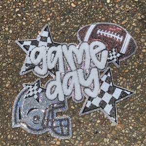 Puede incluir: Un conjunto de parches termoadhesivos con el texto game day en letras de felpa blancas. El diseño incluye un balón de fútbol americano marrón, un casco plateado y dos estrellas con un patrón de cuadros blanco y negro, con lentejuelas brillantes.