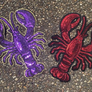 May include: Two lobster-shaped patches featuring dense sequin embroidery. One patch is vibrant purple with a gold-coloured border, while the other is deep red with a black border. Both feature detailed claws, antennae, and segmented tails.