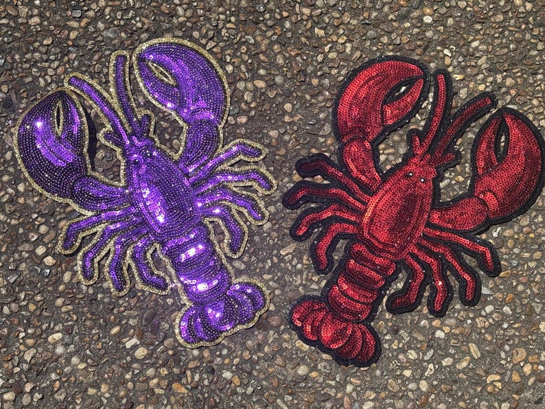 May include: Two lobster-shaped patches featuring dense sequin embroidery. One patch is vibrant purple with a gold-coloured border, while the other is deep red with a black border. Both feature detailed claws, antennae, and segmented tails.
