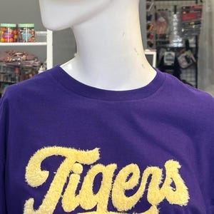 May include: A purple t-shirt with the word "Tigers" in yellow, fuzzy lettering. A gray and tan baseball cap with the word "Tigers" in purple script and a paw print design is on top of the shirt.