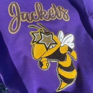 May include: A purple sweatshirt with the word "Jackets" in gold sequined script. The sweatshirt features a cartoon bee wearing star-shaped sunglasses. The bee has yellow and black stripes and white wings. A fun, casual apparel item.