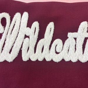 May include: A maroon fabric with the word "Wildcats" in white, textured, cursive lettering. The letters are thick and raised, creating a 3D effect. The background is a solid maroon color.