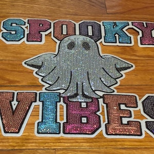Op de afbeelding: Een decoratieve patch met een zilveren paillettengeest gecentreerd tussen de woorden SPOOKY VIBES. De letters zijn gevuld met veelkleurige, glinsterende pailletten in tinten blauw, roze en koper, allemaal omlijnd in wit op een plat oppervlak.