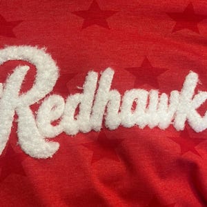 May include: A red fabric garment featuring a repeating pattern of tonal stars across the surface. The word Redhawks is written in large, white, fuzzy, textured script lettering across the center of the fabric, creating a distinct contrast.