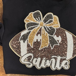 May include: Black sweatshirt with a sequined football design. The football is brown with white stripes and a bow in black, gold, and white. The word "Saints" is written in white script below the football.