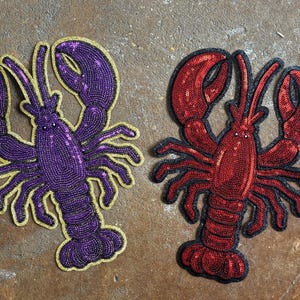 May include: Two lobster-shaped patches featuring sequin embellishments. One patch is purple with a gold-coloured border, and the other patch is red with a black border. Both patches display detailed claws, antennae, and segmented bodies.