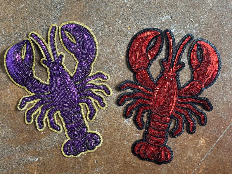 May include: Two lobster-shaped patches featuring sequin embellishments. One patch is purple with a gold-coloured border, and the other patch is red with a black border. Both patches display detailed claws, antennae, and segmented bodies.