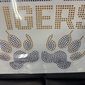 Op de afbeelding: Witte stof met het woord "TIGERS" geschreven in gouden strass steentjes. Daaronder staan ​​pootafdrukken en druppelvormen, eveneens gemaakt van gouden en paarse strass steentjes. Het ontwerp staat op een zwart oppervlak.