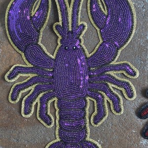 May include: A decorative lobster patch featuring purple sequins and beads arranged in a detailed crustacean shape. The design is outlined with a shimmering gold border, highlighting the two large claws, multiple legs, and segmented tail structure.