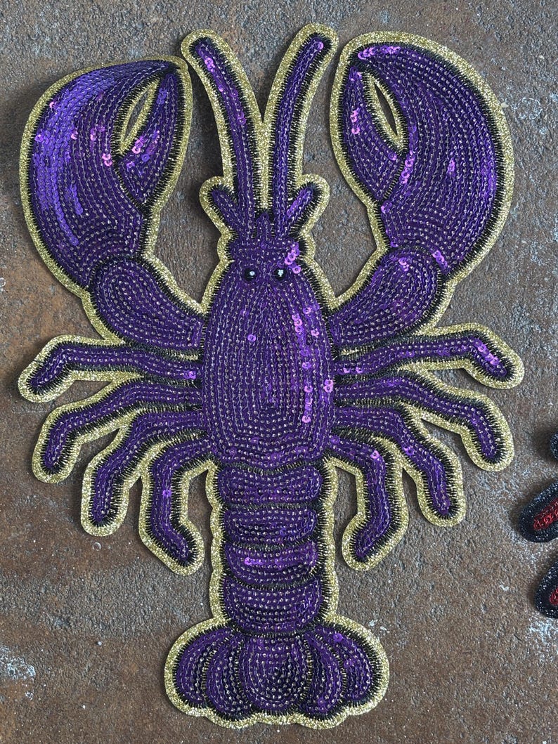 May include: A decorative lobster patch featuring purple sequins and beads arranged in a detailed crustacean shape. The design is outlined with a shimmering gold border, highlighting the two large claws, multiple legs, and segmented tail structure.