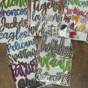 May include: A collection of assorted sports-themed iron-on patches packaged in clear plastic sleeves. The patches feature various team names like Lions, Broncos, Tigers, and Wildcats, alongside footballs, helmets, and bows in diverse colours and textures.