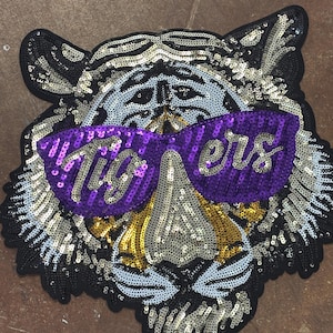 May include: A tiger head patch crafted from black, white, and gold sequins. The tiger wears purple sequined sunglasses with the word "Tigers" written in white script across the lenses. The design features detailed facial markings and black borders.