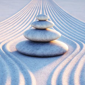 May include: A stack of smooth, gray stones balanced on top of each other in a zen garden setting. The stones are arranged in a pyramid shape on a bed of white sand with raked lines.