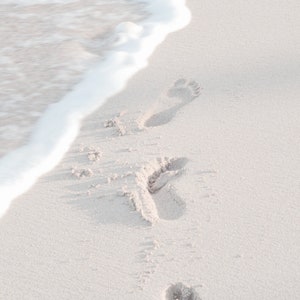 May include: Footprints in the sand leading towards the ocean waves. The white sand is wet from the receding tide.