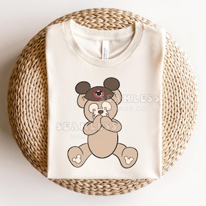 Può includere: T-shirt color crema con un personaggio di orso dei cartoni animati con un cappello marrone e un disegno a occhio rosso e bianco. L'orso è seduto con le mani che coprono la bocca. La maglietta è su una superficie tessuta.