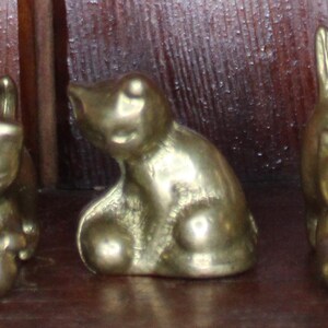 May include: Three brass cat figurines in various poses. The cats are golden in color and appear to be made of solid metal. One cat is sitting, another is standing, and the third is in a playful position.