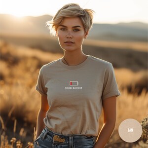 Op de afbeelding: Een beige T-shirt met een witte afbeelding van een bijna lege batterij en de tekst "Social Battery".