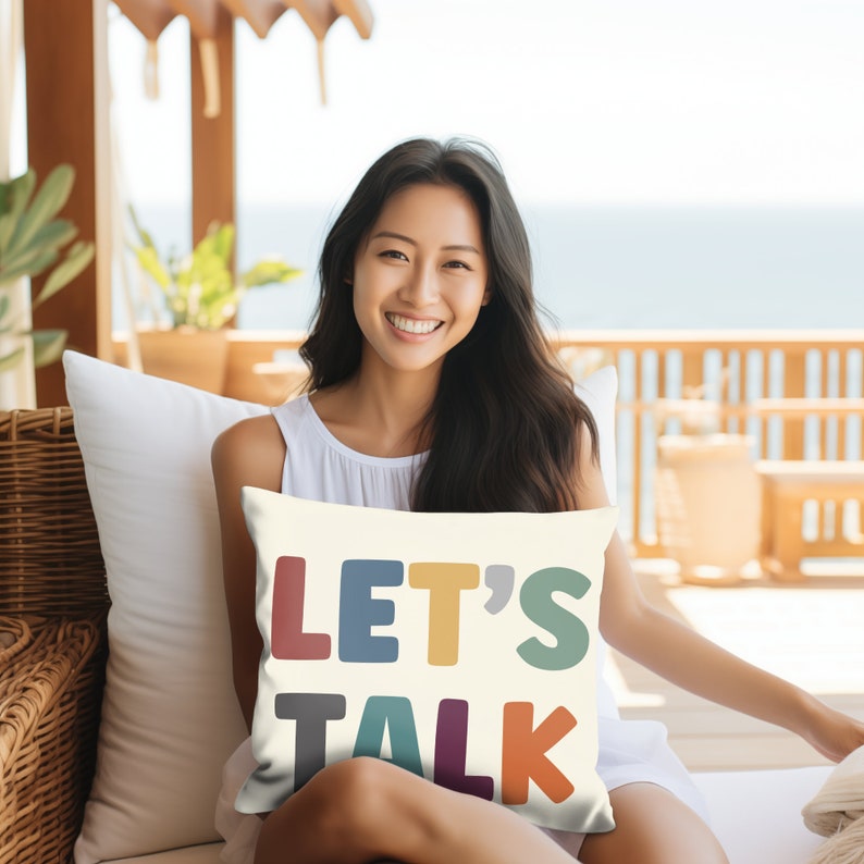 May include: A woman is sitting on a wicker chair holding a white pillow with the words "LET'S TALK" in colorful letters. The pillow is a decorative accent for a home.