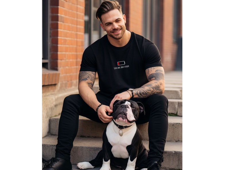Op de afbeelding: Een man draagt een zwart T-shirt met de tekst "Social Battery" en een pictogram voor een lege batterij. Hij zit op een trap met een zwart-witte bulldog.