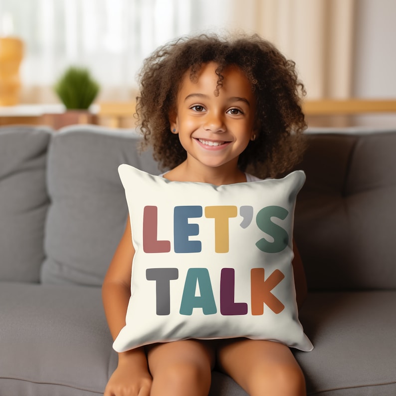 May include: A white throw pillow with the words "LET'S TALK" in colorful, block letters. The pillow is being held by a young girl with curly hair, sitting on a gray couch.