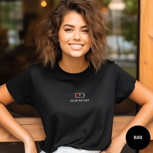 Op de afbeelding: Een zwart T-shirt met een witte afbeelding van een batterij met de tekst "SOCIAL BATTERY".