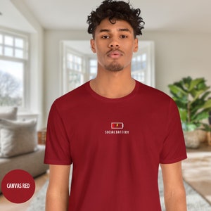 Op de afbeelding: Een rood T-shirt met een witte afbeelding van een batterij met een bliksem en de tekst "SOCIAL BATTERY".