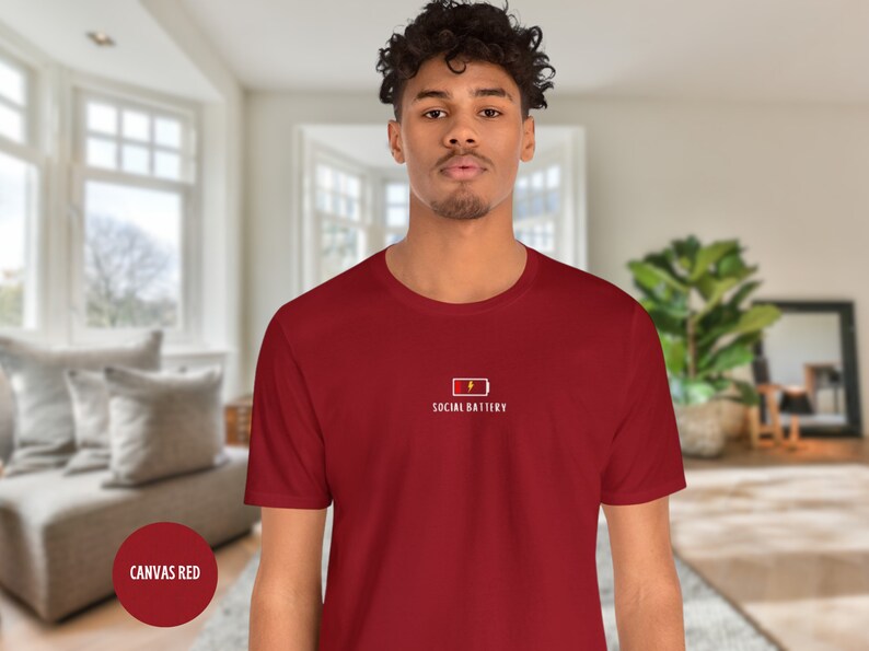 Op de afbeelding: Een rood T-shirt met een witte afbeelding van een batterij met een bliksem en de tekst "SOCIAL BATTERY".