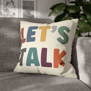 May include: A cream-colored throw pillow with the text "LET'S TALK" in colorful, bold letters. The letters are in shades of blue, yellow, green, purple, and orange.