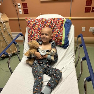 Puede incluir: Una persona en pijama descansa en una cama de hospital, sosteniendo dos osos de peluche. El pijama es azul oscuro con un estampado de veh&iacute;culos. Una almohada con un dise&ntilde;o de c&oacute;mic est&aacute; detr&aacute;s de la persona.