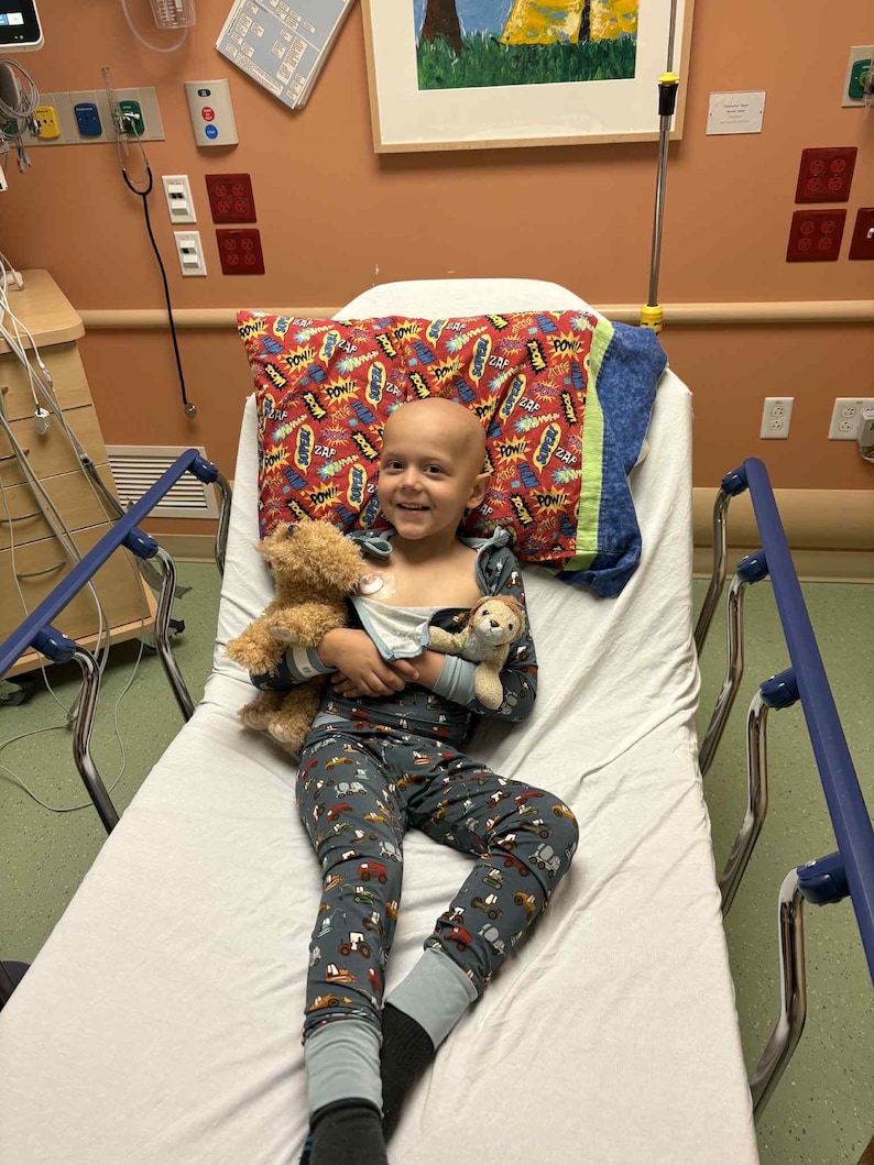 Puede incluir: Una persona en pijama descansa en una cama de hospital, sosteniendo dos osos de peluche. El pijama es azul oscuro con un estampado de veh&iacute;culos. Una almohada con un dise&ntilde;o de c&oacute;mic est&aacute; detr&aacute;s de la persona.