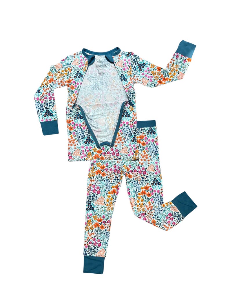 K&ouml;nnte beinhalten: Ein zweiteiliger Schlafanzug mit floralem Muster in Orange, Rosa, Blau und Gr&uuml;n auf wei&szlig;em Grund. Das lang&auml;rmlige Oberteil hat einen Rei&szlig;verschluss vorne und einen tealfarbenen Besatz. Die passende Hose hat tealfarbene B&uuml;ndchen.