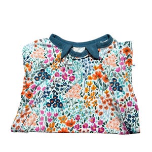 K&ouml;nnte beinhalten: Gefaltetes wei&szlig;es Kleidungsst&uuml;ck mit floralem Muster und teal-farbenem Kragen. Das Blumenmuster umfasst orange, rosa und blaue Blumen mit gr&uuml;nen Bl&auml;ttern. Das Kleidungsst&uuml;ck ist sauber gefaltet.