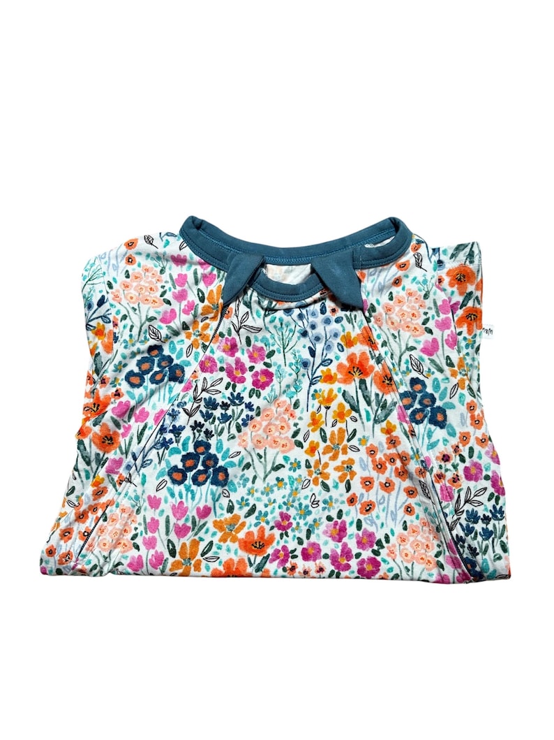 K&ouml;nnte beinhalten: Gefaltetes wei&szlig;es Kleidungsst&uuml;ck mit floralem Muster und teal-farbenem Kragen. Das Blumenmuster umfasst orange, rosa und blaue Blumen mit gr&uuml;nen Bl&auml;ttern. Das Kleidungsst&uuml;ck ist sauber gefaltet.