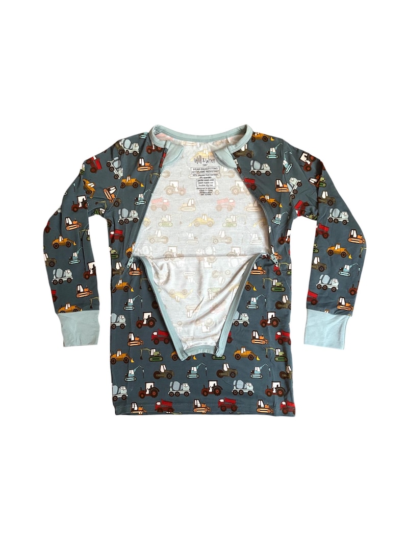 Puede incluir: Un conjunto de pijama de manga larga en color verde azulado apagado, estampado con varios veh&iacute;culos de construcci&oacute;n en tonos amarillos, naranjas, rojos y grises. Los pu&ntilde;os y el cuello son de color azul claro. El pijama tiene una cremallera completa.