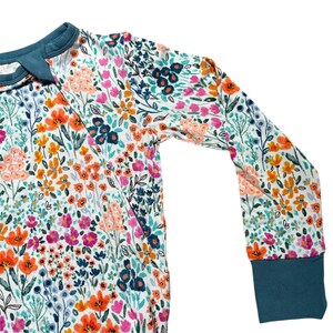 K&ouml;nnte beinhalten: Lang&auml;rmliges Kleidungsst&uuml;ck mit einem lebendigen Blumenmuster. Das Design zeigt einen wei&szlig;en Hintergrund, der mit orangefarbenen, rosa und blauen Blumen verziert ist. Die Manschetten und der Kragen sind einfarbig t&uuml;rkis. Das Kleidungsst&uuml;ck ist aus einem weichen, bequemen Material.