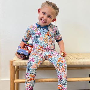 K&ouml;nnte beinhalten: Ein Kinder-Schlafanzug-Set mit floralem Muster, bestehend aus einem kurz&auml;rmeligen Oberteil und einer Hose. Der Schlafanzug hat ein farbenfrohes Blumenmuster in Rosa-, Orange-, Blau- und Lilat&ouml;nen. Das Set hat einen t&uuml;rkisfarbenen Besatz. Das Kind h&auml;lt einen Spielzeug-LKW.