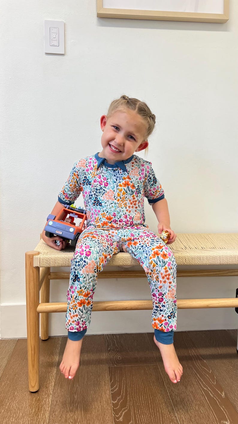 K&ouml;nnte beinhalten: Ein Kinder-Schlafanzug-Set mit floralem Muster, bestehend aus einem kurz&auml;rmeligen Oberteil und einer Hose. Der Schlafanzug hat ein farbenfrohes Blumenmuster in Rosa-, Orange-, Blau- und Lilat&ouml;nen. Das Set hat einen t&uuml;rkisfarbenen Besatz. Das Kind h&auml;lt einen Spielzeug-LKW.