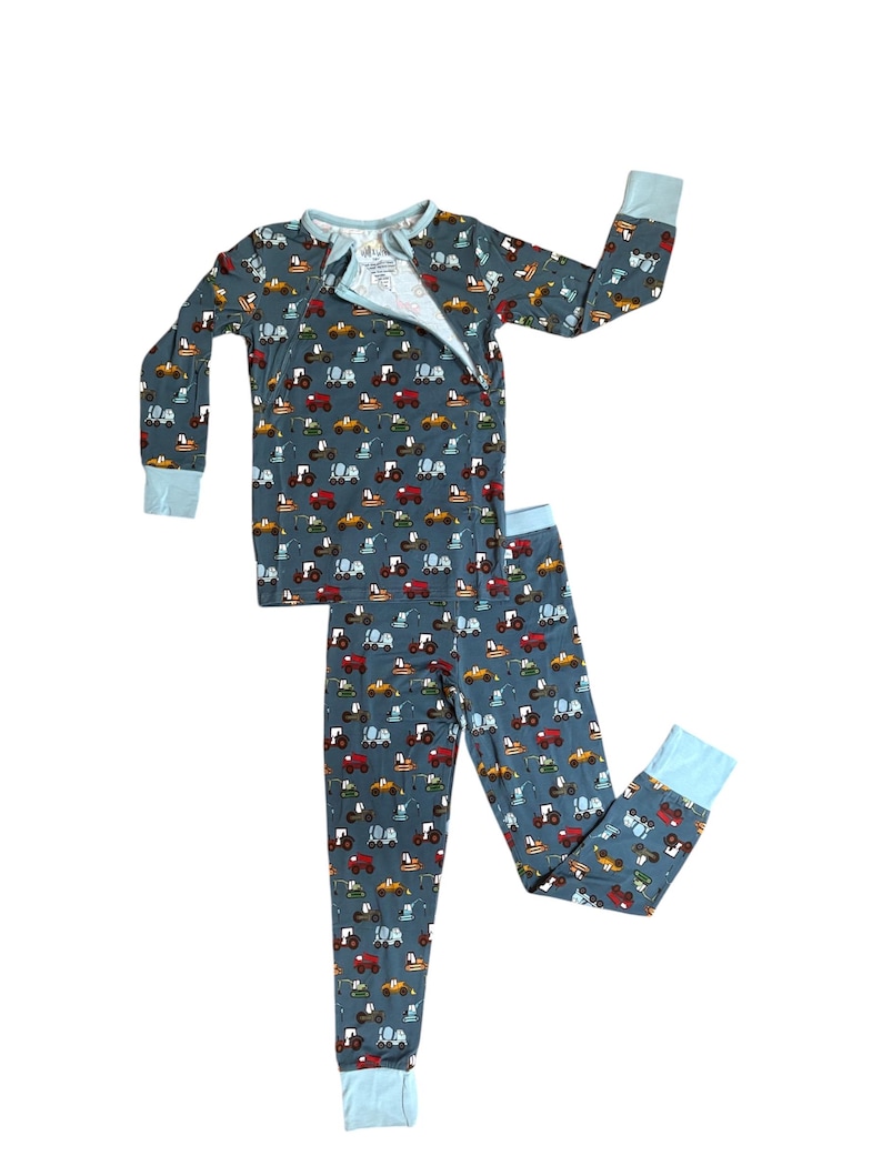 Puede incluir: Un conjunto de pijama de dos piezas en color azul gris&aacute;ceo, con un estampado repetido de tractores coloridos. La parte superior de manga larga tiene cierre de cremallera y pu&ntilde;os azul claro. Los pantalones a juego tambi&eacute;n tienen pu&ntilde;os azul claro.