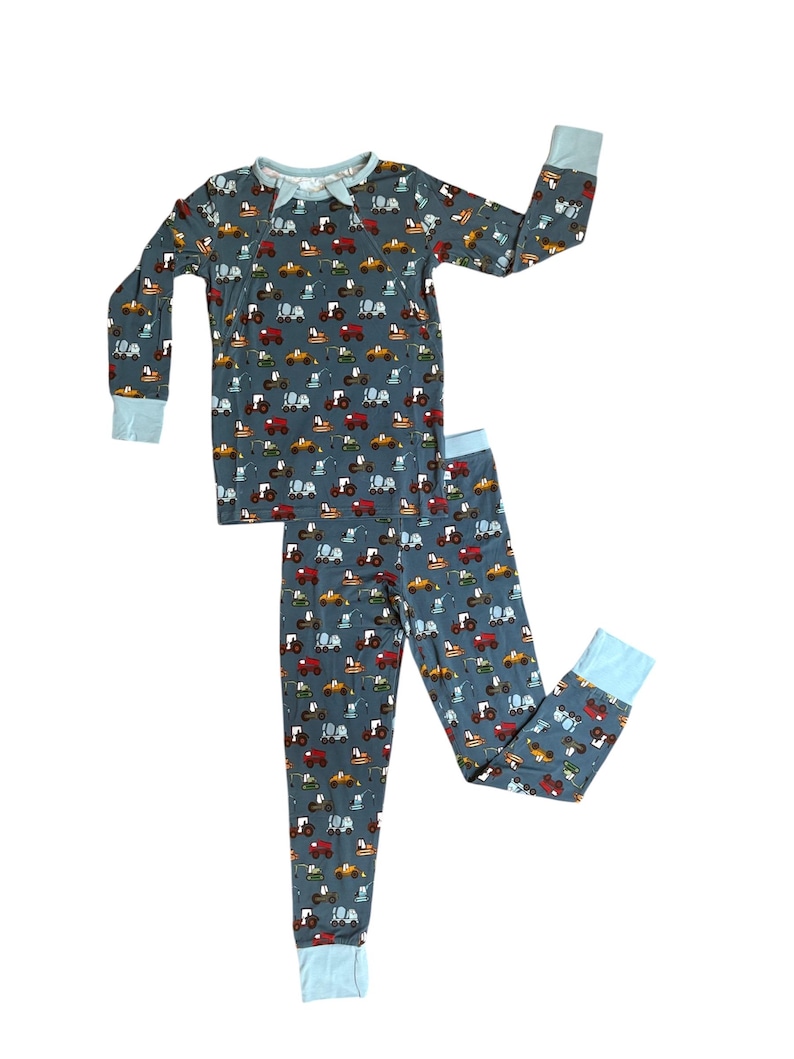 Puede incluir: Un conjunto de pijama de dos piezas con un estampado de tractores coloridos sobre un fondo verde azulado oscuro. La parte superior de manga larga y los pantalones tienen pu&ntilde;os y ribetes de color azul claro. El pijama est&aacute; dise&ntilde;ado para la comodidad y el sue&ntilde;o.