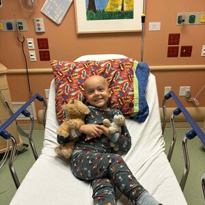 Puede incluir: Un ni&ntilde;o sonr&iacute;e en una cama de hospital, con un pijama estampado y sosteniendo dos peluches. La cama tiene una almohada colorida. La habitaci&oacute;n incluye equipo m&eacute;dico y una pintura enmarcada.