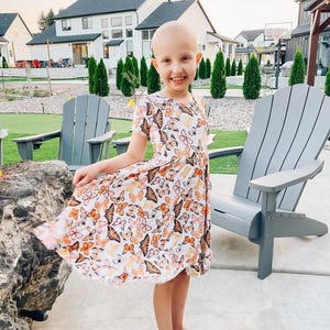 May include: A young girl with short, shaved hair wearing a white dress with a butterfly print. The dress has a flowing skirt and short sleeves. The girl is standing in front of a house with a green lawn.