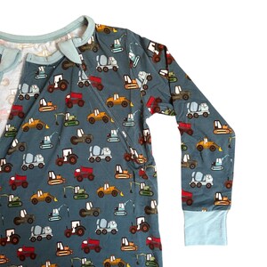 Puede incluir: Mono de manga larga de color verde azulado con un estampado repetido de veh&iacute;culos de construcci&oacute;n coloridos. El mono tiene un ribete azul claro alrededor del cuello, la cremallera y los pu&ntilde;os. Los veh&iacute;culos incluyen camiones, excavadoras y hormigoneras.