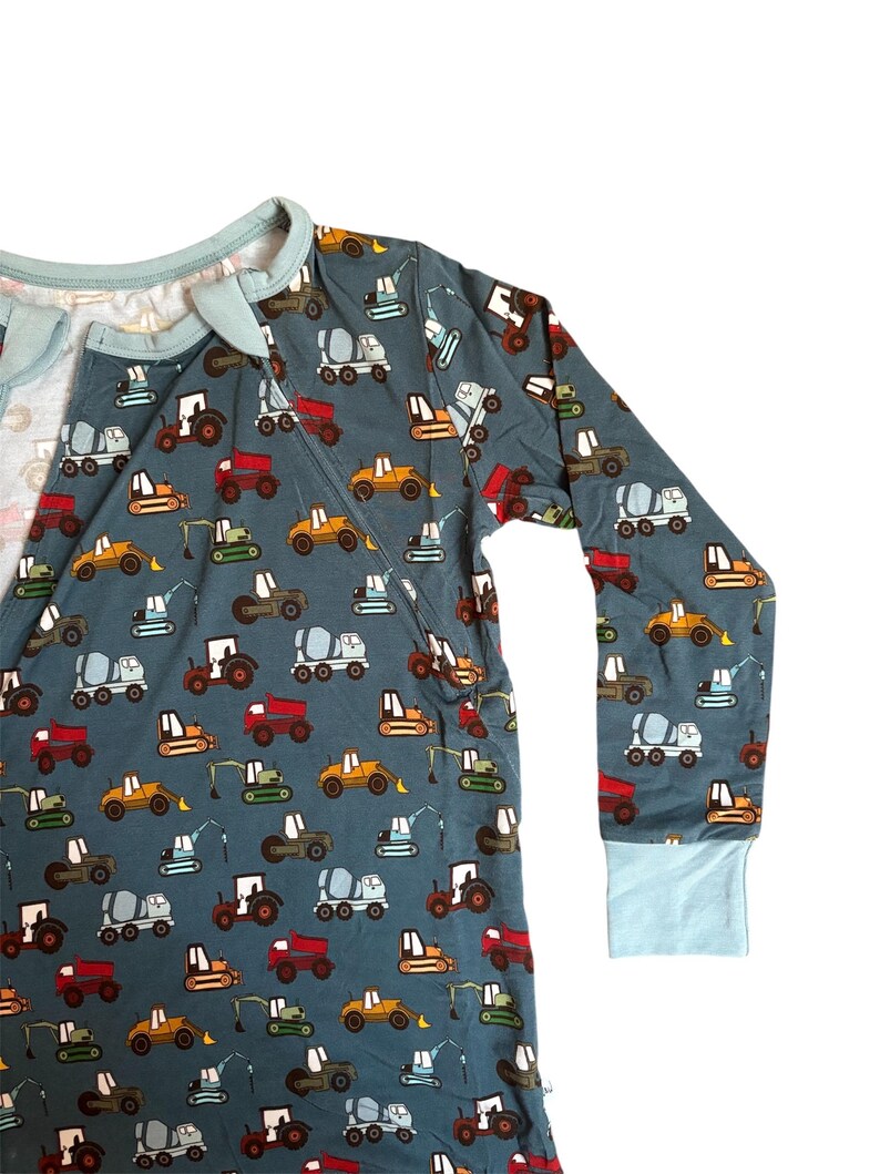 Puede incluir: Mono de manga larga de color verde azulado con un estampado repetido de veh&iacute;culos de construcci&oacute;n coloridos. El mono tiene un ribete azul claro alrededor del cuello, la cremallera y los pu&ntilde;os. Los veh&iacute;culos incluyen camiones, excavadoras y hormigoneras.