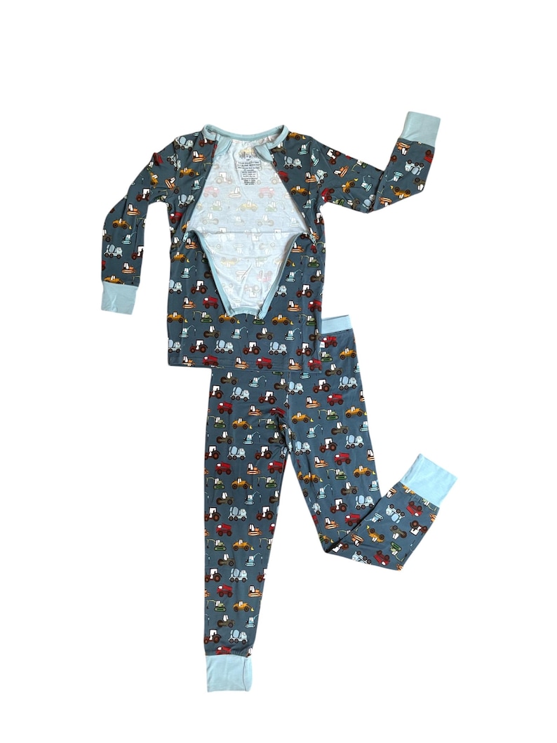 Puede incluir: Un conjunto de pijama de dos piezas en un color azul gris&aacute;ceo apagado, estampado con tractores coloridos. La parte superior de manga larga y los pantalones tienen pu&ntilde;os y ribetes de color azul claro. La parte superior tiene una abertura parcial.