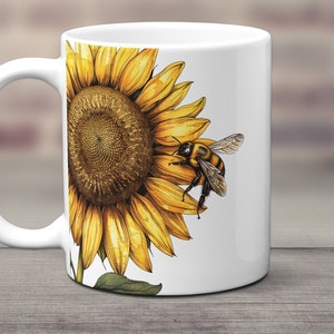 May include: White ceramic mug with a sunflower and a bee design. The sunflower is yellow with a brown center and green leaves. The bee is yellow and black with wings.