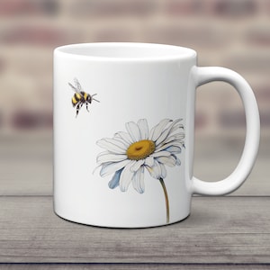 May include: White ceramic mug with a graphic of a bumblebee flying towards a white daisy with a yellow center.