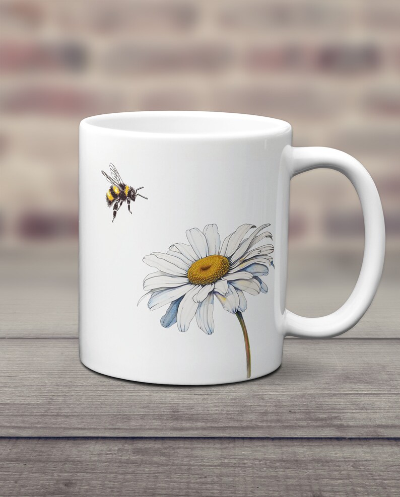 May include: White ceramic mug with a graphic of a bumblebee flying towards a white daisy with a yellow center.
