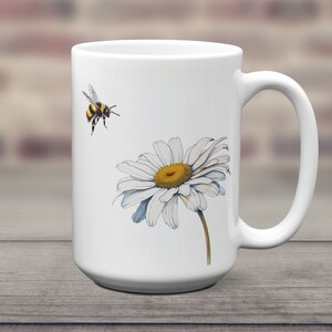 May include: White ceramic mug with a graphic of a bumblebee flying towards a white daisy with a yellow center.