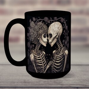 May include: Black coffee mug with a graphic of two skeletons embracing, surrounded by roses. The skeletons are wearing floral crowns and have intricate details.