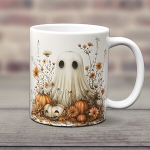 K&ouml;nnte beinhalten: Wei&szlig;e Keramiktasse mit einer Geisterillustration, umgeben von orangefarbenen K&uuml;rbissen und Herbstlaub. Der Geist hat zwei schwarze Augen und einen wei&szlig;en, lakenartigen K&ouml;rper.
